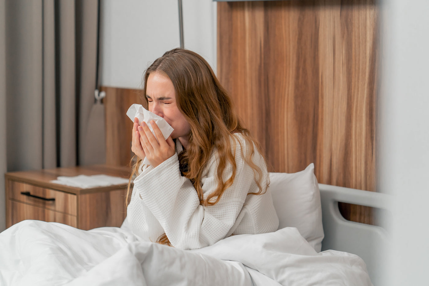 Vrouw in badjas niest in een tissue terwijl ze in bed zit, mogelijk door huisstofmijtallergie.